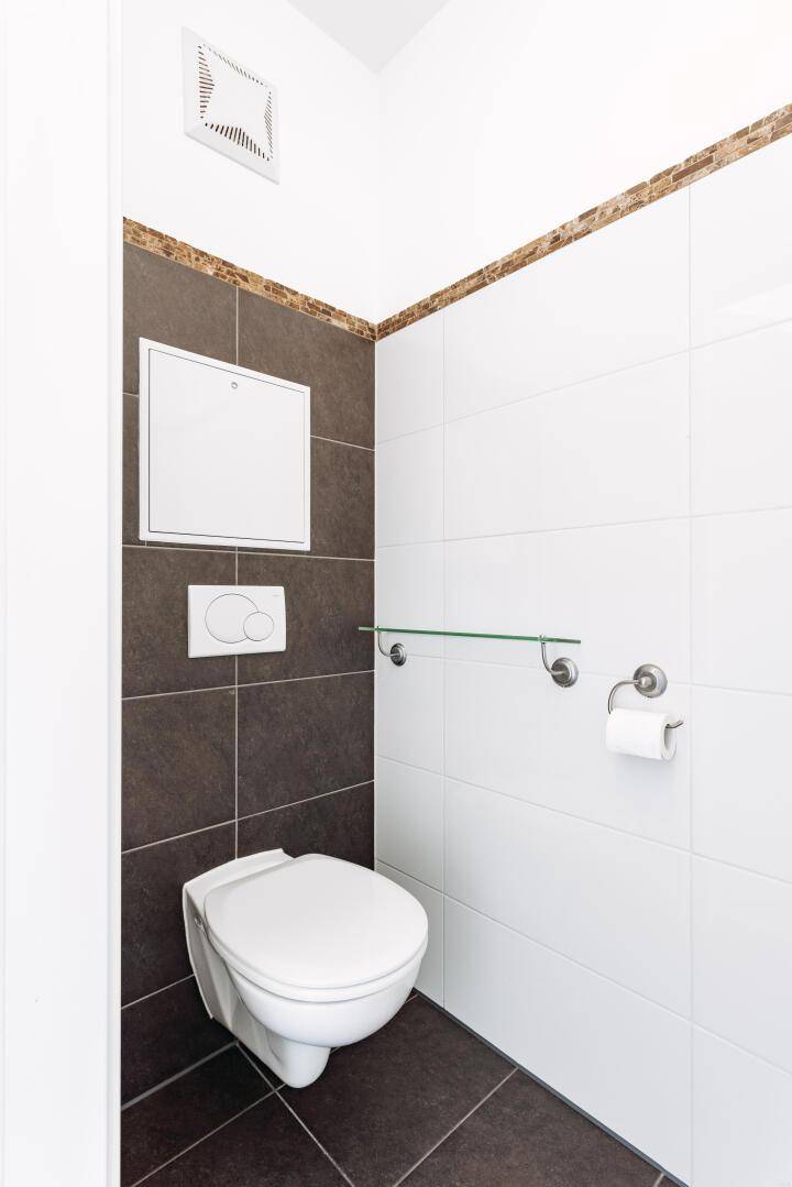 Separate Toilette mit Wand-WC, Fliesen an Wänden und Boden sowie einer Lüftung.