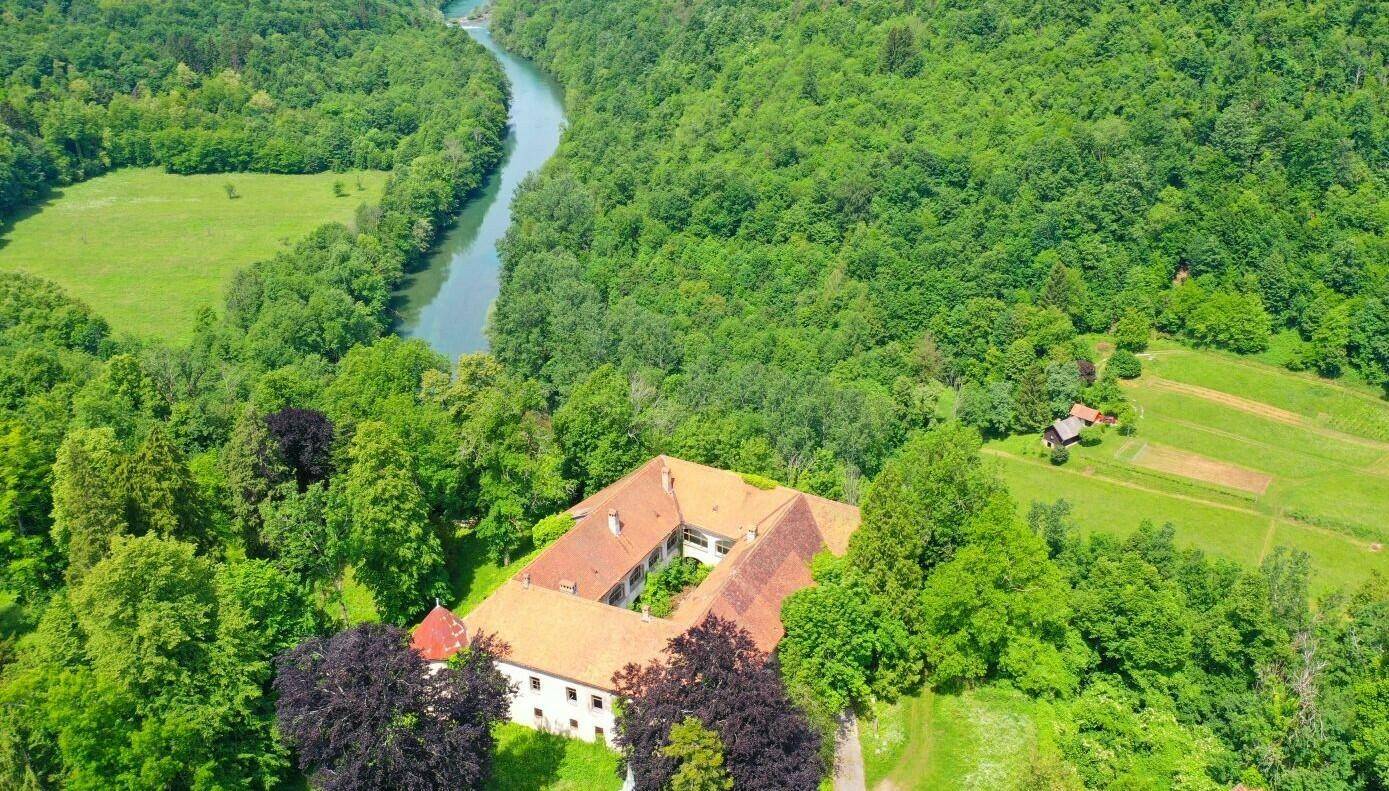 Historisches Gebäude mit rotem Dach, umgeben von dichtem Wald und einem Fluss in einer weiten Landschaft.
