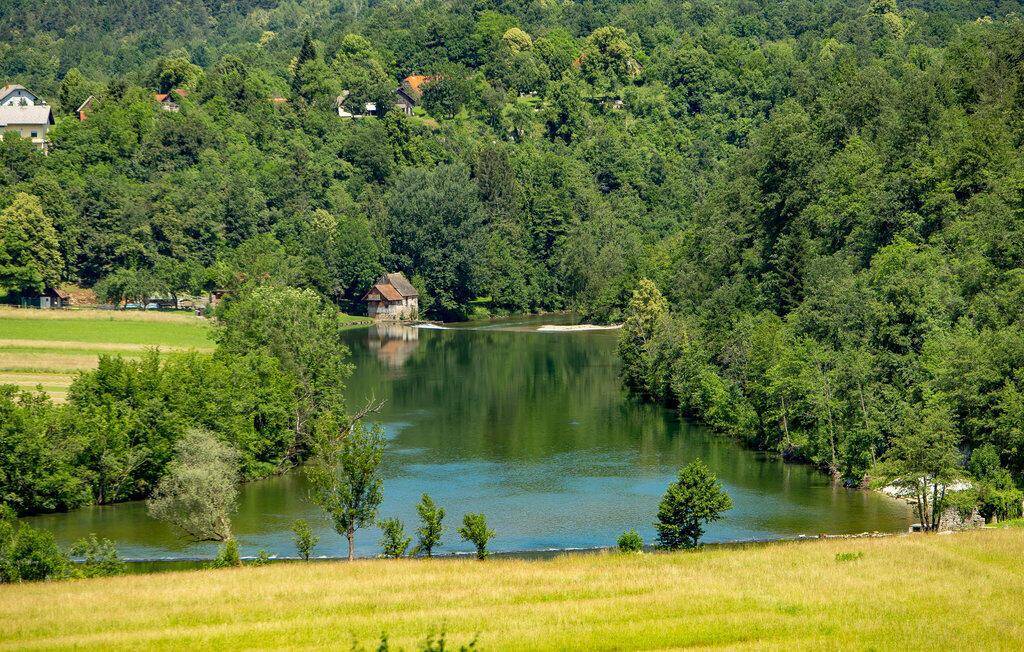 Idyllischer See mit einem kleinen Haus am Ufer, umgeben von üppiger grüner Natur und bewaldeten Hügeln.