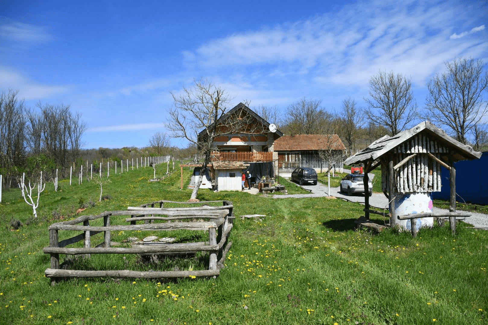 Haus mit Balkon, einem Brunnen und Holzzaun in einer grünen Umgebung mit geparkten Autos.