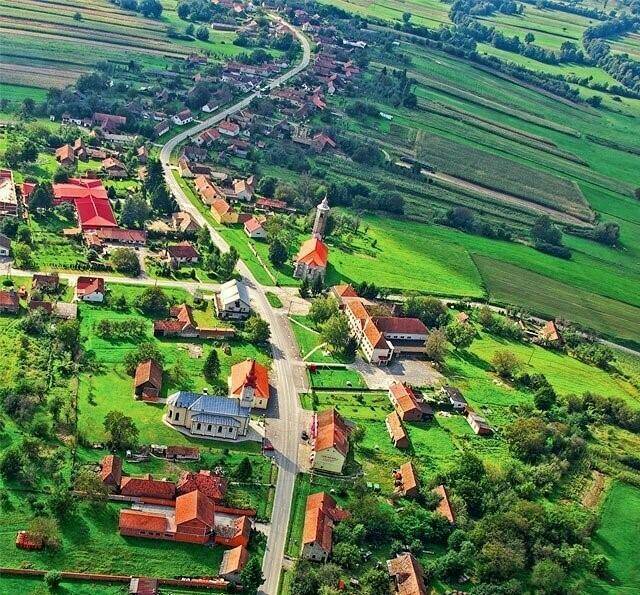 Luftaufnahme eines kleinen Dorfes eingebettet in eine grüne, hügelige Landschaft mit Häusern und Straßen.