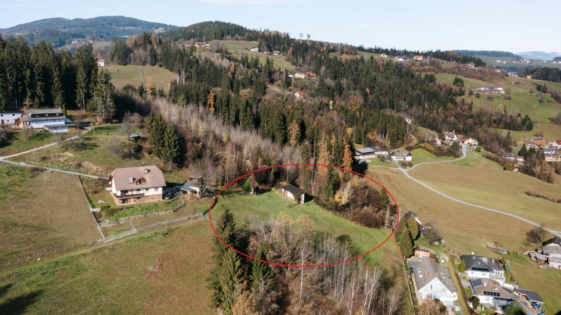 Luftaufnahme einer hügeligen Landschaft mit verstreuten Häusern, dichten Wäldern und grünen Wiesen unter blauem Himmel.