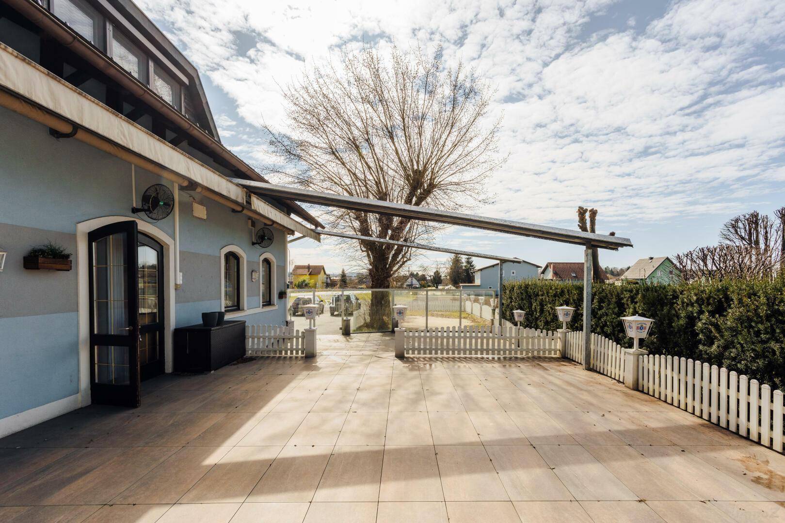 Große gepflasterte Terrasse mit Markise, umzäunt von einem weißen Lattenzaun und Bäumen.