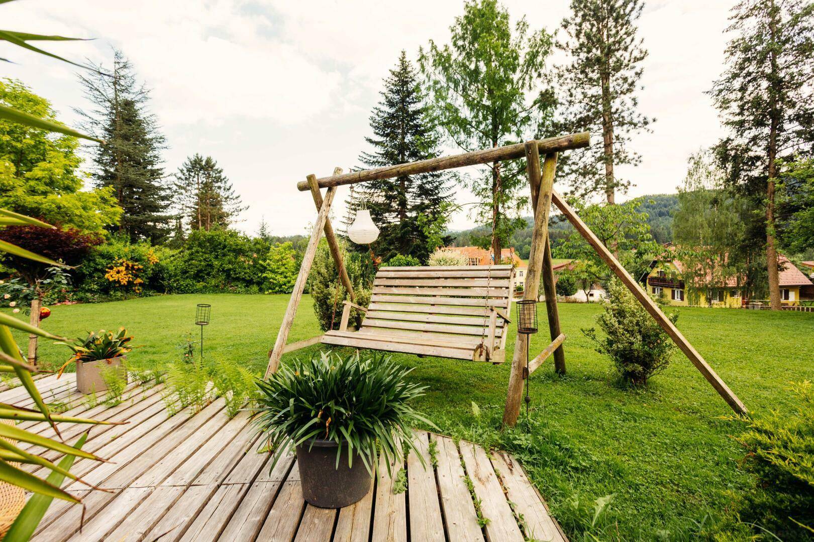 Rustikale Holzschaukel auf einer Holzterrasse, umgeben von einem gepflegten Rasen und Bäumen.