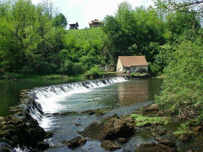 Rustikales Steinhaus mit rotem Ziegeldach am Flussufer, neben einem Wasserfall und umgeben von grünen Hügeln.