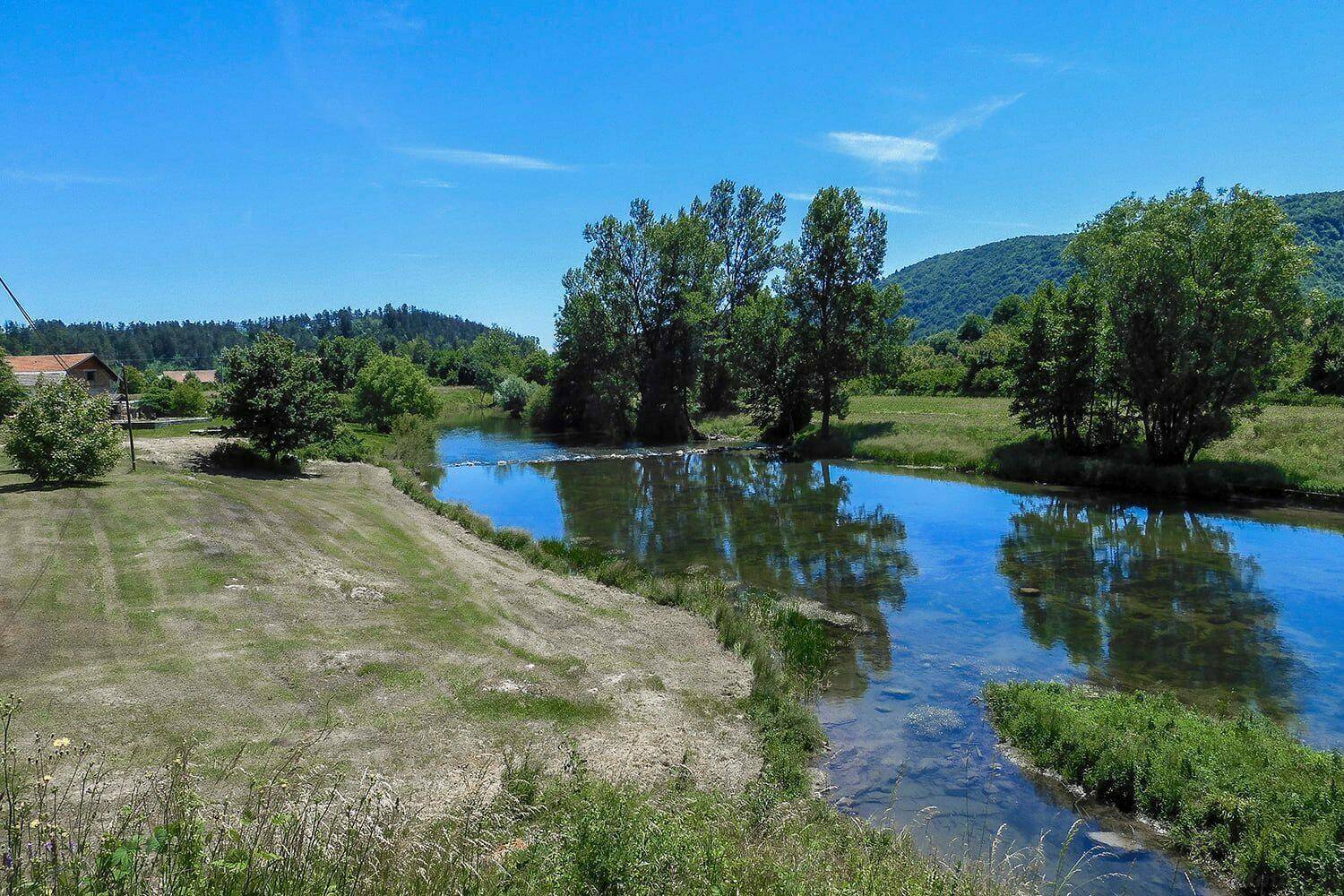 Flusslauf durch grüne Wiesen und Bäume, mit Häusern und Hügeln in der Ferne unter klarem Himmel.