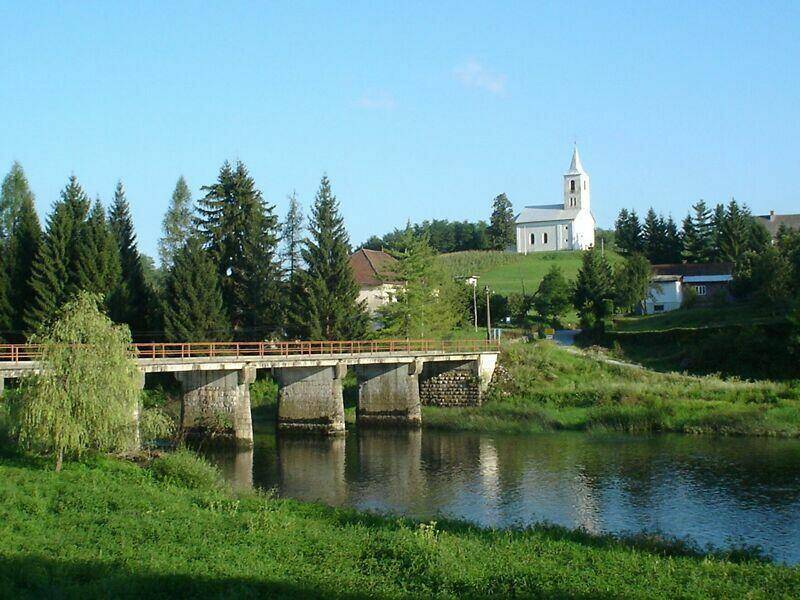 Historische Brücke über einen Fluss mit einer Kirche auf einem Hügel im Hintergrund, umgeben von Natur.
