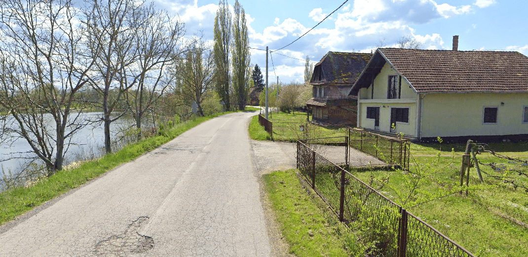 Ländliche Straße entlang eines Sees mit Häusern und Bäumen im Hintergrund.