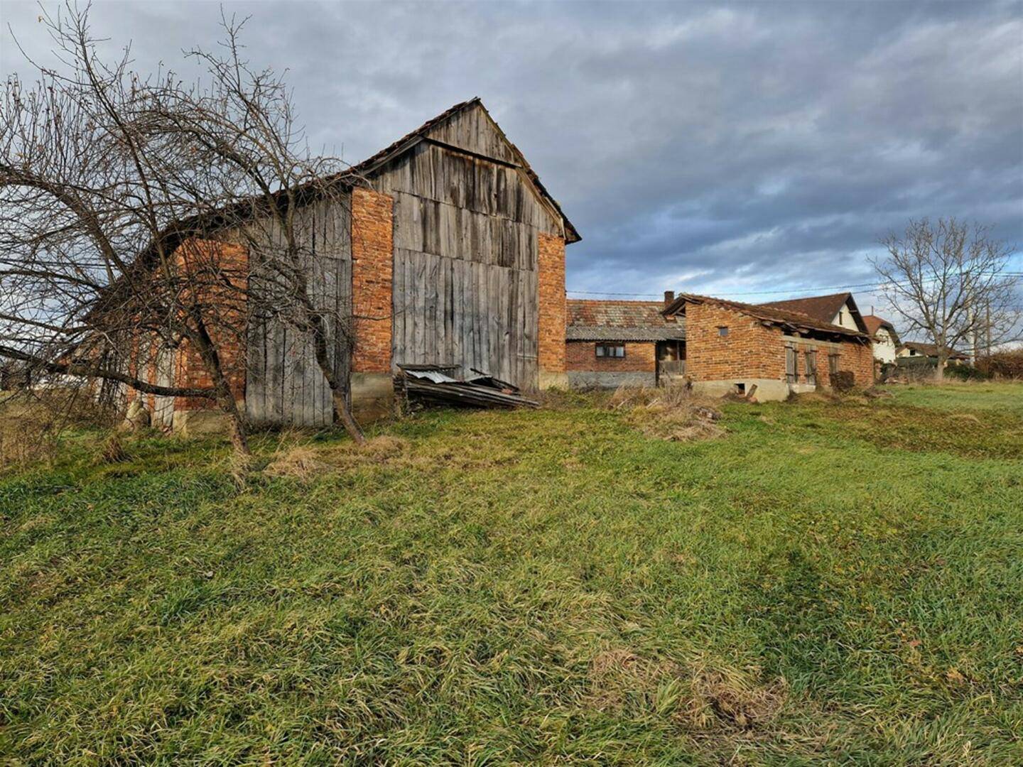 Große alte Scheune aus Backstein und Holz mit schrägem Dach, umgeben von Gras unter bewölktem Himmel.