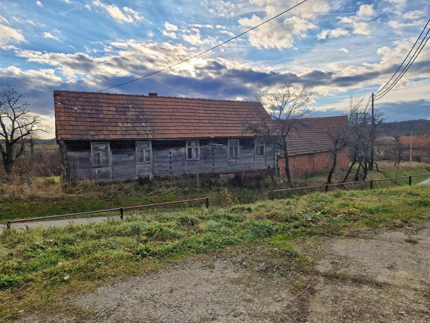 Altes, verfallenes Holzhaus mit Ziegeldach in ländlicher Umgebung unter einem bewölkten Himmel.
