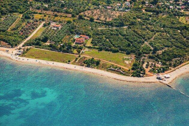 Luftaufnahme einer Küstenlinie mit Sandstrand, türkisblauem Meer und Gebäuden inmitten grüner Landschaft.