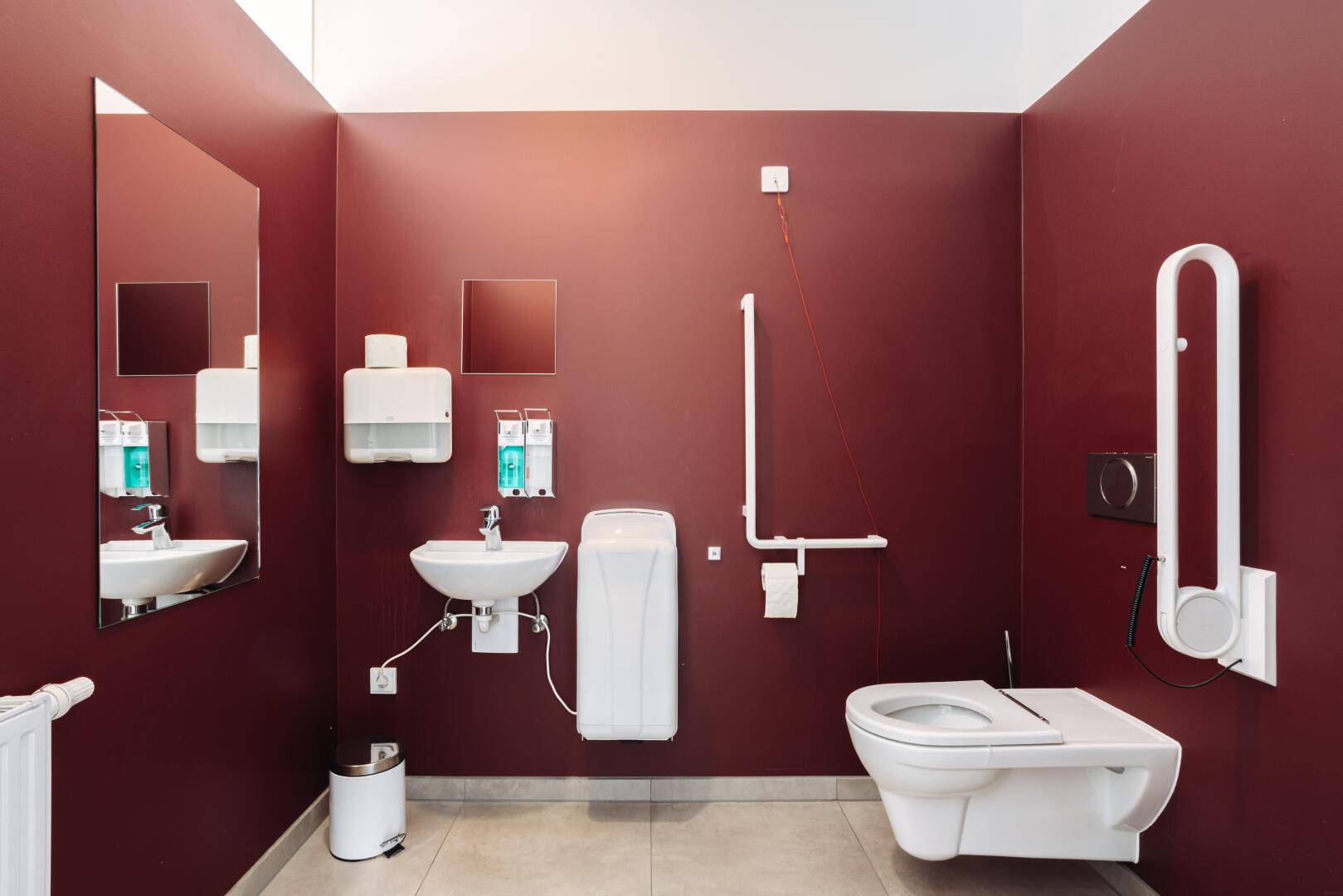 Barrierefreie Toilette mit Haltegriffen, Waschbecken und roter Wandfarbe.