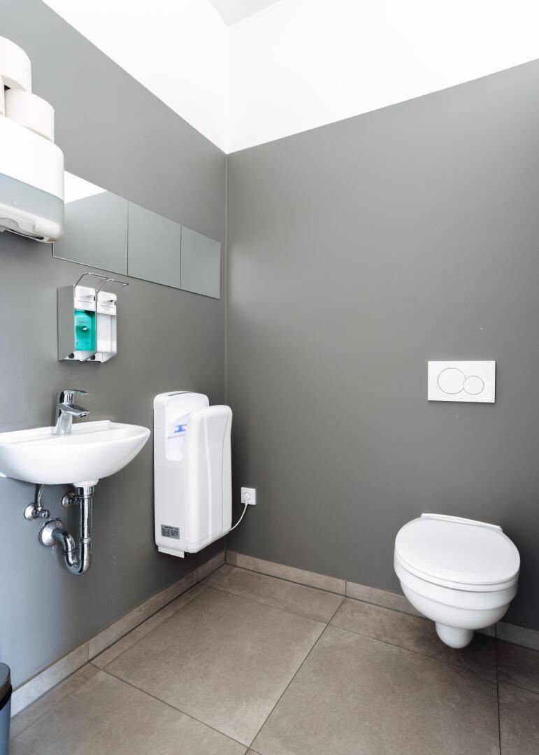 Toilette mit Waschbecken, Handtrockner und grauen Fliesen an Wänden und Boden.