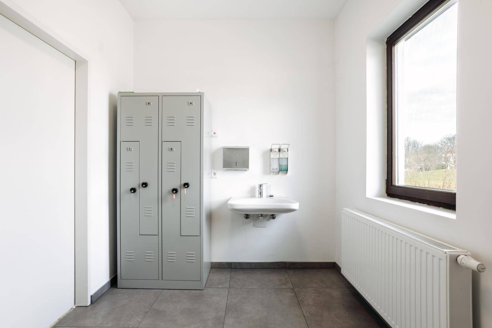 Umkleideraum mit Spinden, Waschbecken, Fliesenboden und einem Fenster.