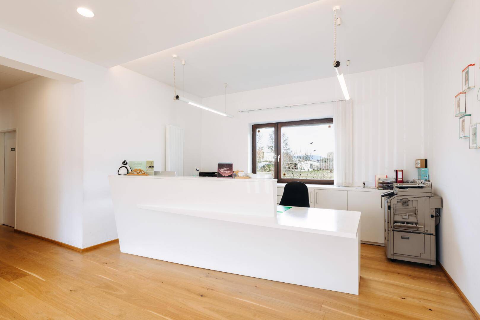Moderner Empfangsbereich mit weißer Rezeptionstheke, Holzfußboden und großem Fenster.