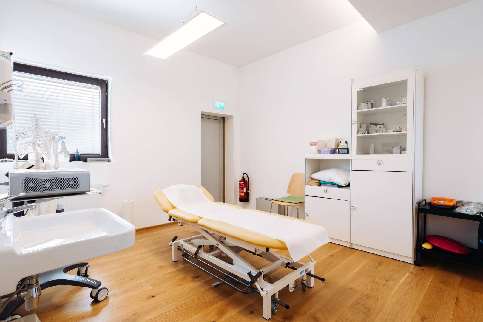 Therapieraum mit Behandlungsliege, Waschbecken, Schrank und Holzfußboden.