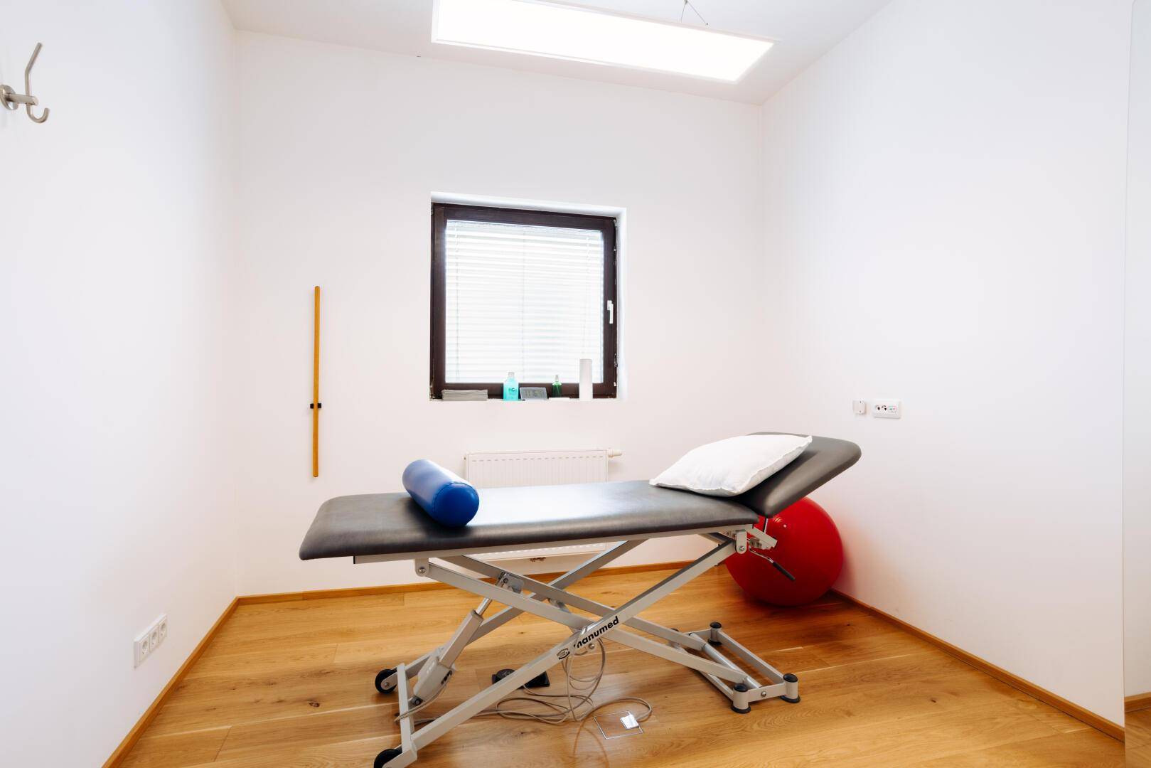 Therapieraum mit einer Behandlungsliege, Holzfußboden und einem Fenster mit Jalousien.