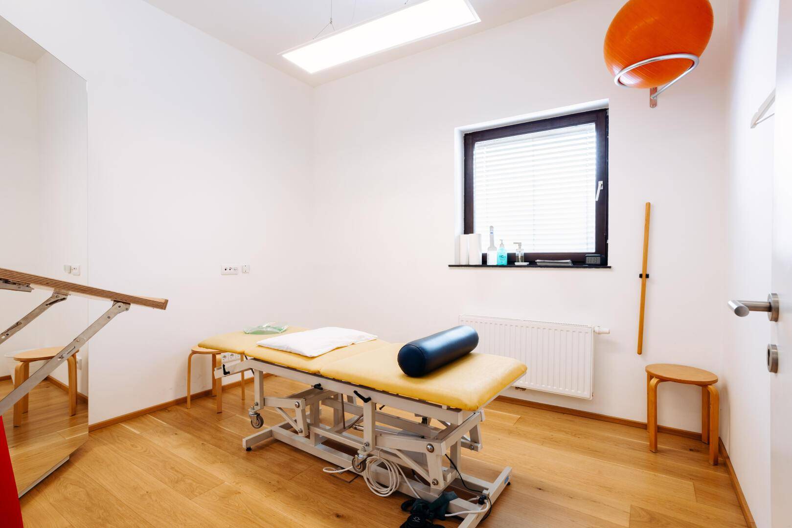 Therapieraum mit einer Behandlungsliege, Holzfußboden und einem Fenster mit Jalousien.