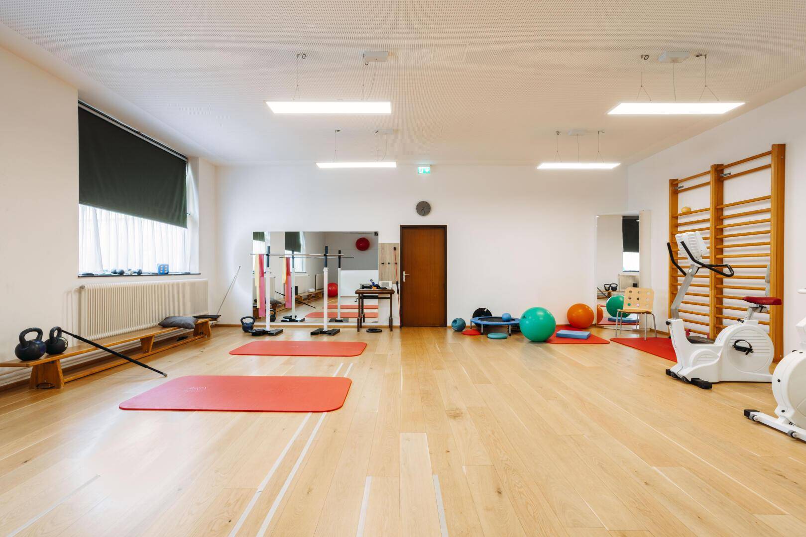 Geräumiger Trainingsraum mit Holzboden, Fitnessgeräten, Spiegeln und Sprossenwänden.