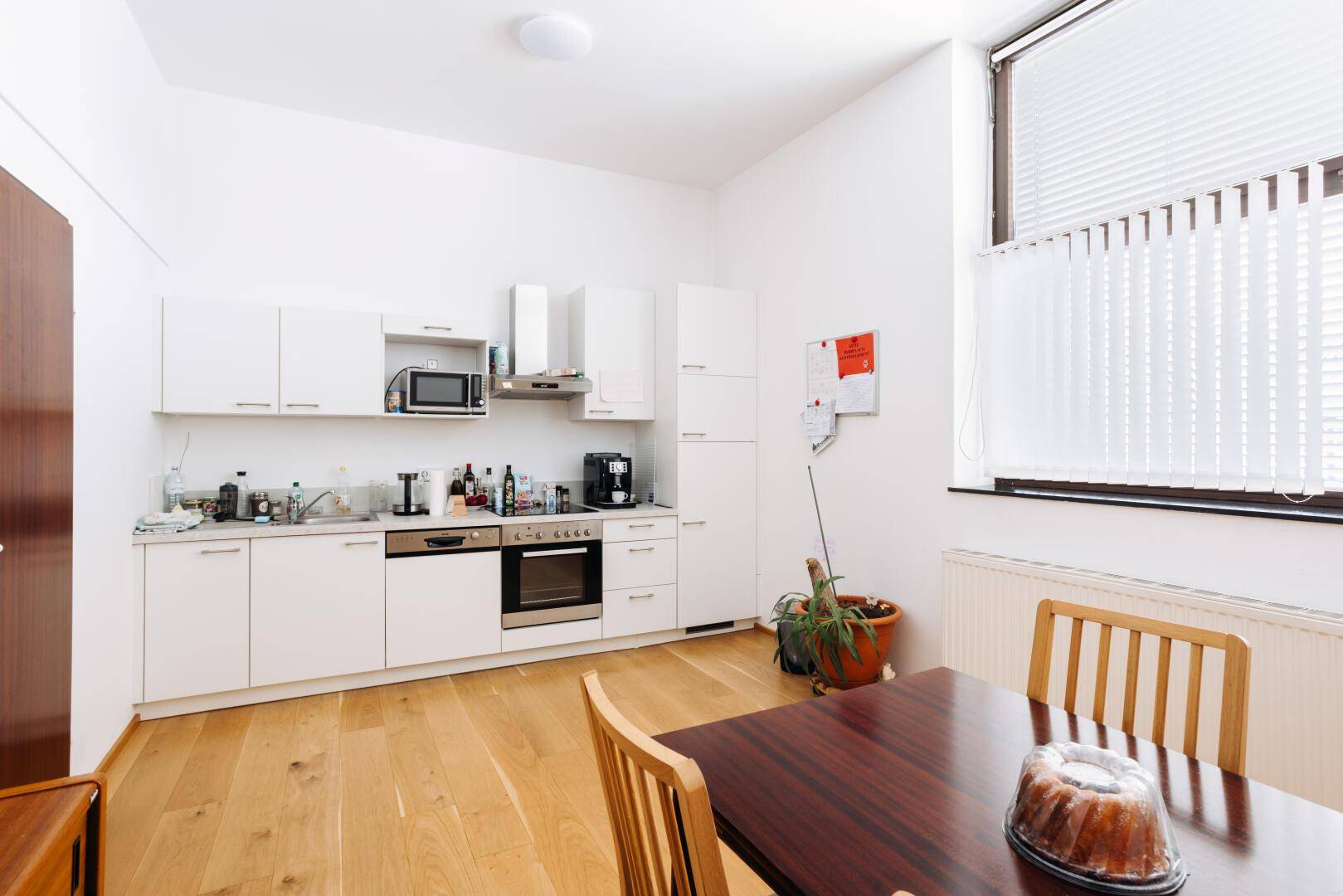 Helle Küche mit weißen Schränken, Holzboden, Esstisch und Fenster mit Jalousien.