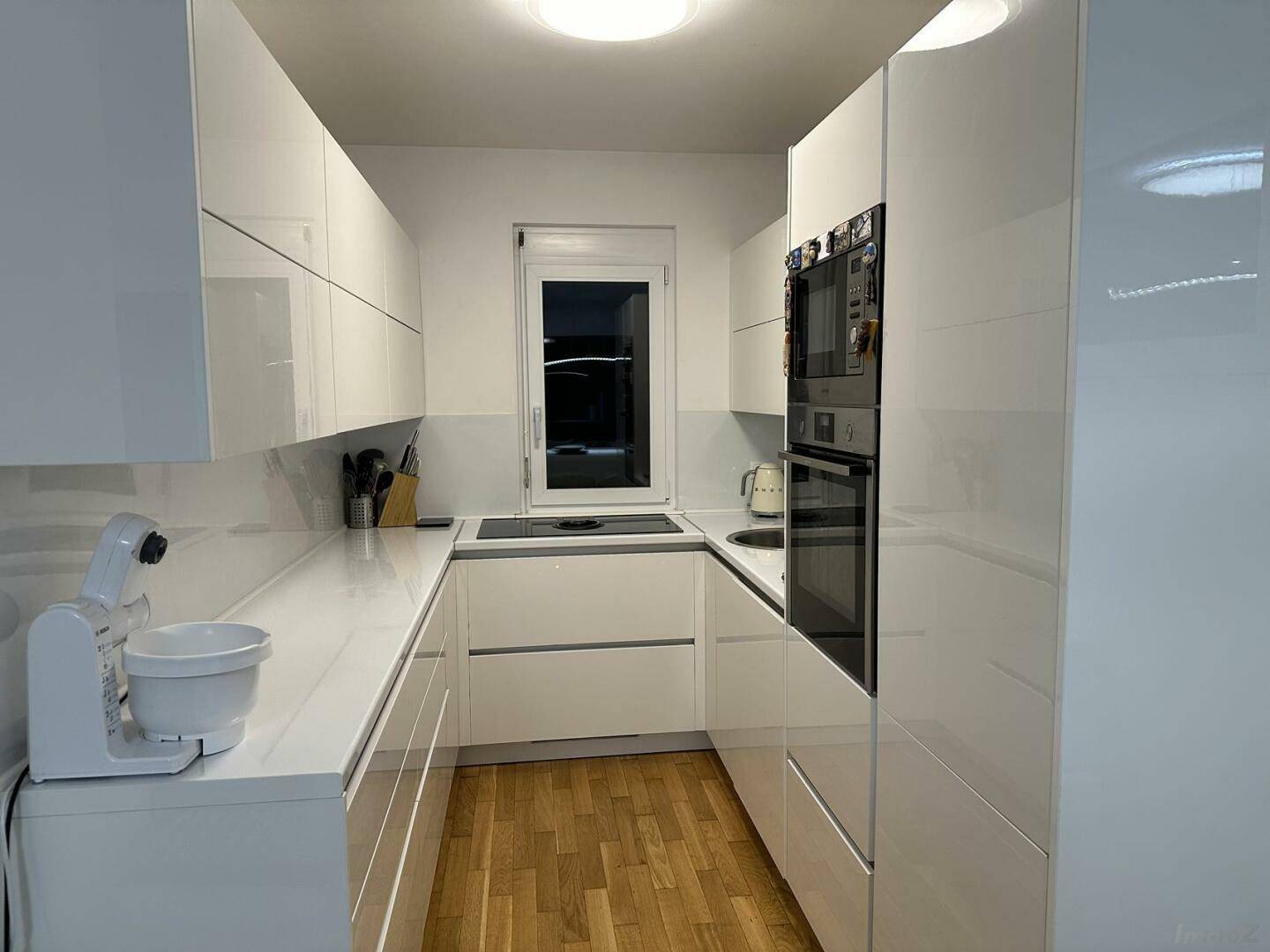 Moderne weiße Küche mit Hochglanzfronten, Einbaugeräten und Fenster für natürliches Licht.