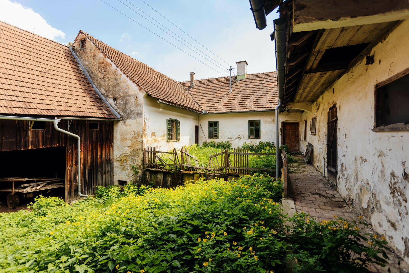 Blick auf den Innenhof eines Bauernhofs mit üppiger Vegetation und den historischen Gebäuden im Hintergrund.
