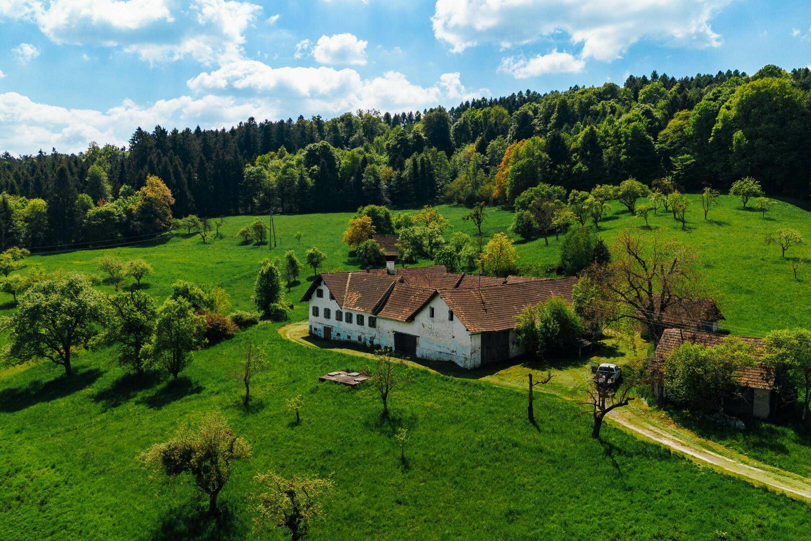 Luftaufnahme eines Bauernhofs in idyllischer ländlicher Umgebung, umgeben von sanften Hügeln und Wäldern.