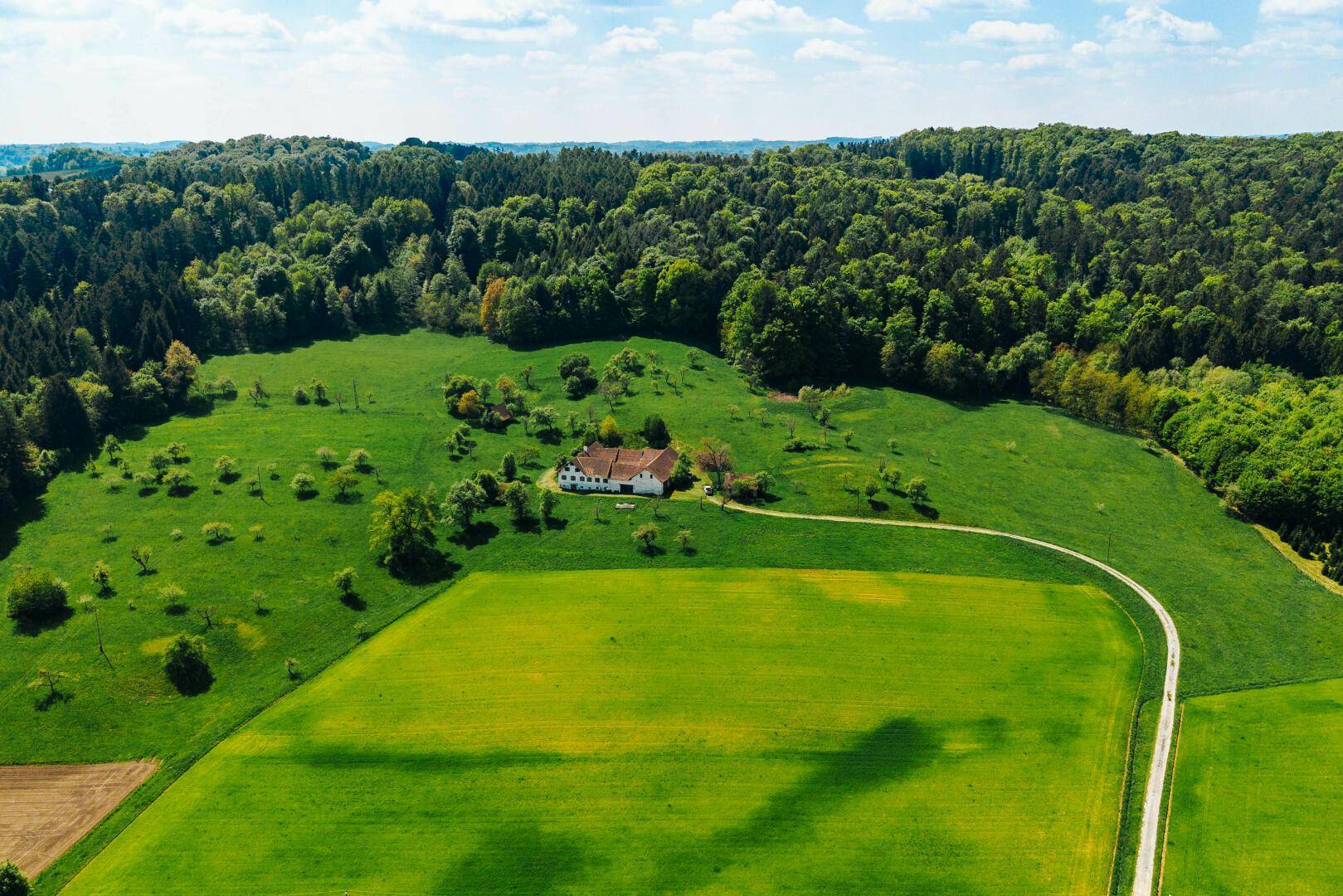 Weitläufige Luftaufnahme eines Bauernhofs in hügeliger Landschaft, umgeben von Feldern und dichtem Wald.