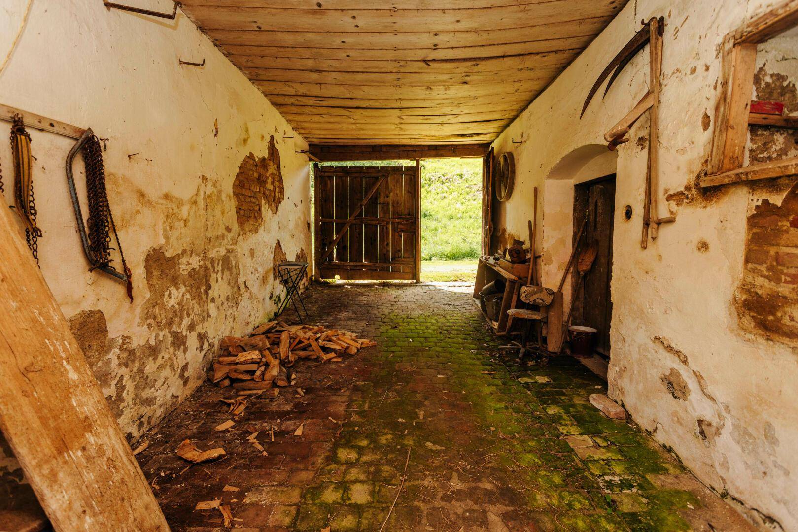 Langer, schmaler Durchgang mit Ziegelboden und Holzbalkendecke, der zu einem offenen Tor führt.