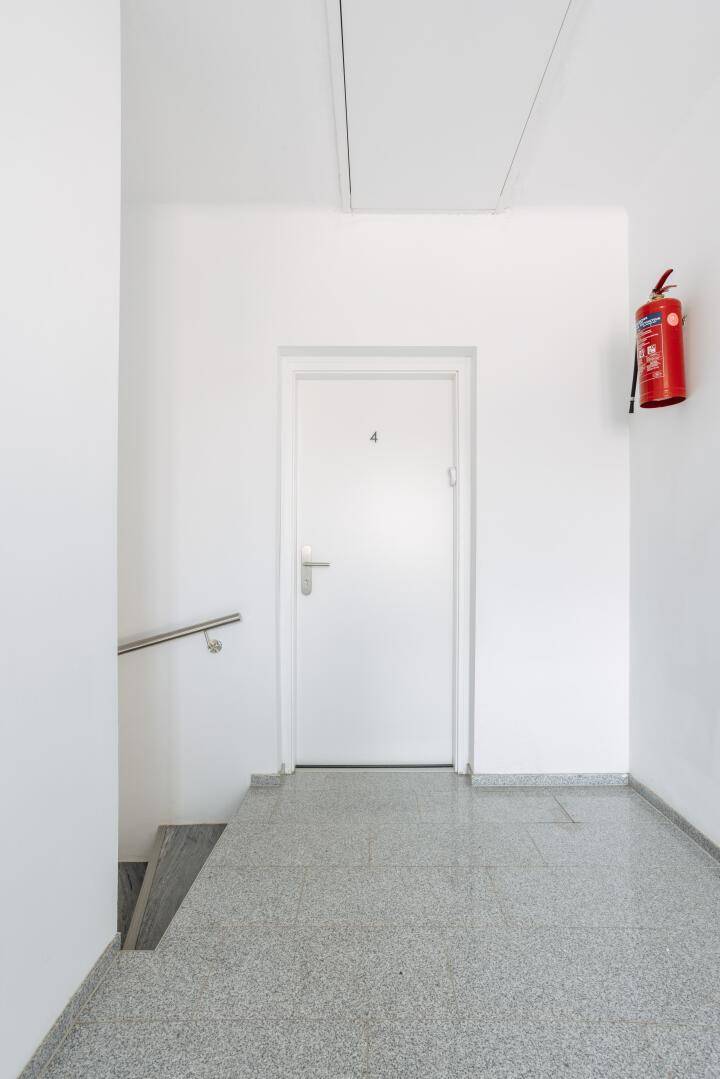 Heller Treppenhausbereich mit weißer Tür, Handlauf und grauem Terrazzoboden.