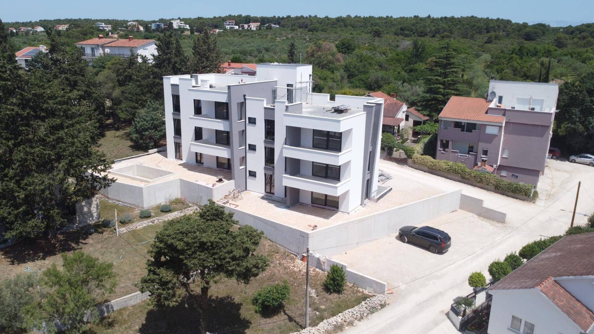 Blick von oben auf das moderne Apartmentgebäude im Bau mit unfertigem Poolbereich und geparktem Auto.