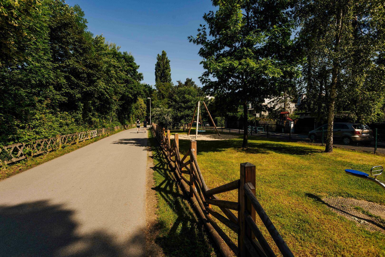 Spazierweg entlang eines Spielplatzes und einer grünen Hecke an einem sonnigen Tag.