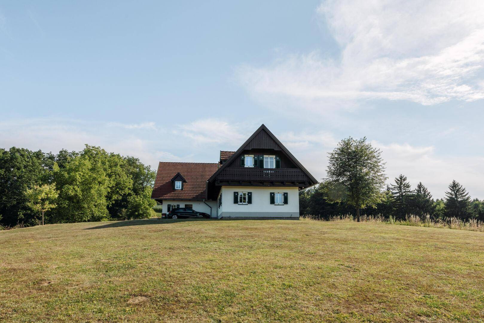 Traditionelles Landhaus mit weißer Fassade, grünen Fensterläden und Satteldach auf einer Anhöhe.