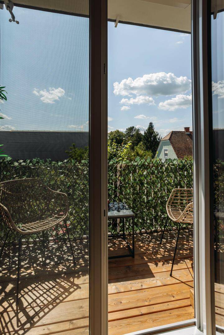 Sonniger Balkon mit Holzboden, Sitzgelegenheiten und grüner Bepflanzung.