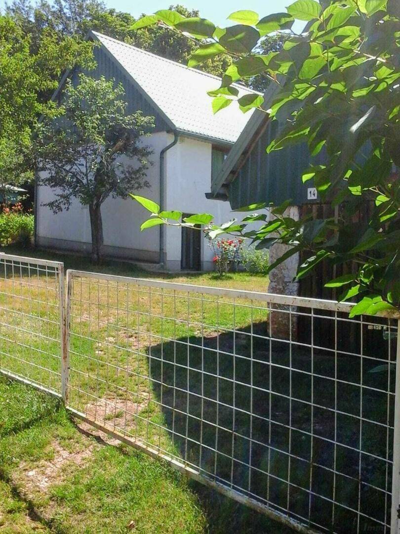 Weißes Haus mit grünem Dach, sichtbar durch einen Metallzaun, umgeben von Bäumen und Gras.