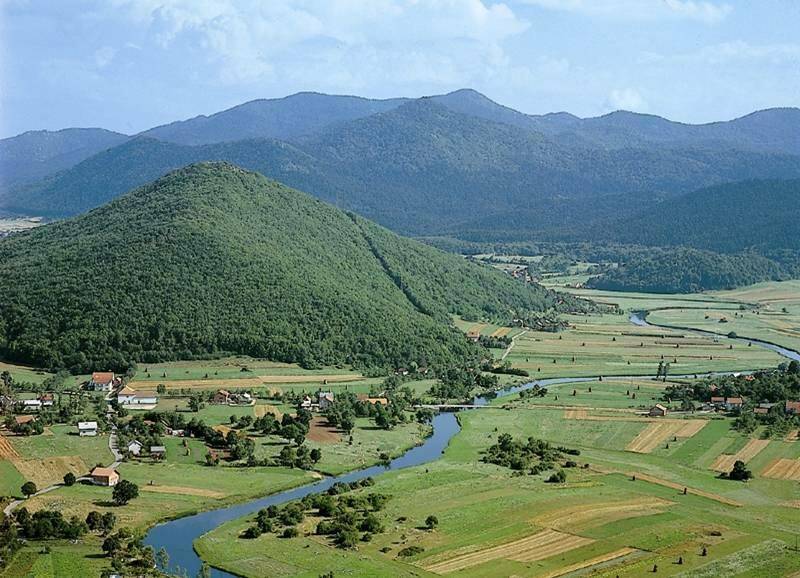 Panoramablick über eine grüne Flusslandschaft mit einem Dorf und bewaldeten Bergen im Hintergrund.