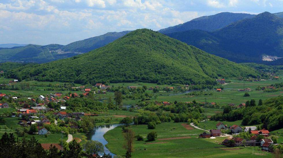 Weitläufige Landschaft mit grünen Hügeln, einem Fluss und einem kleinen Dorf unter blauem Himmel.