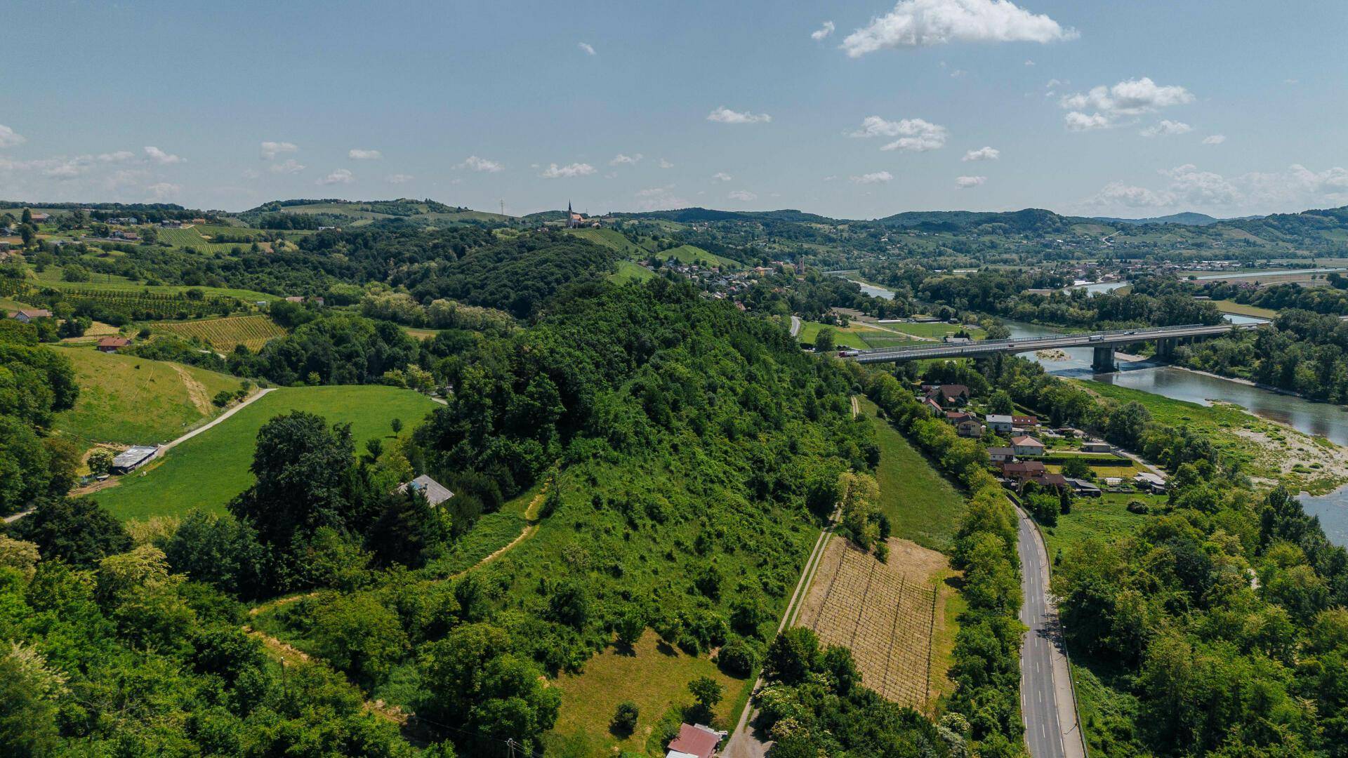 Luftaufnahme einer weitläufigen grünen Hügellandschaft mit Fluss, Straße und vereinzelten Häusern.