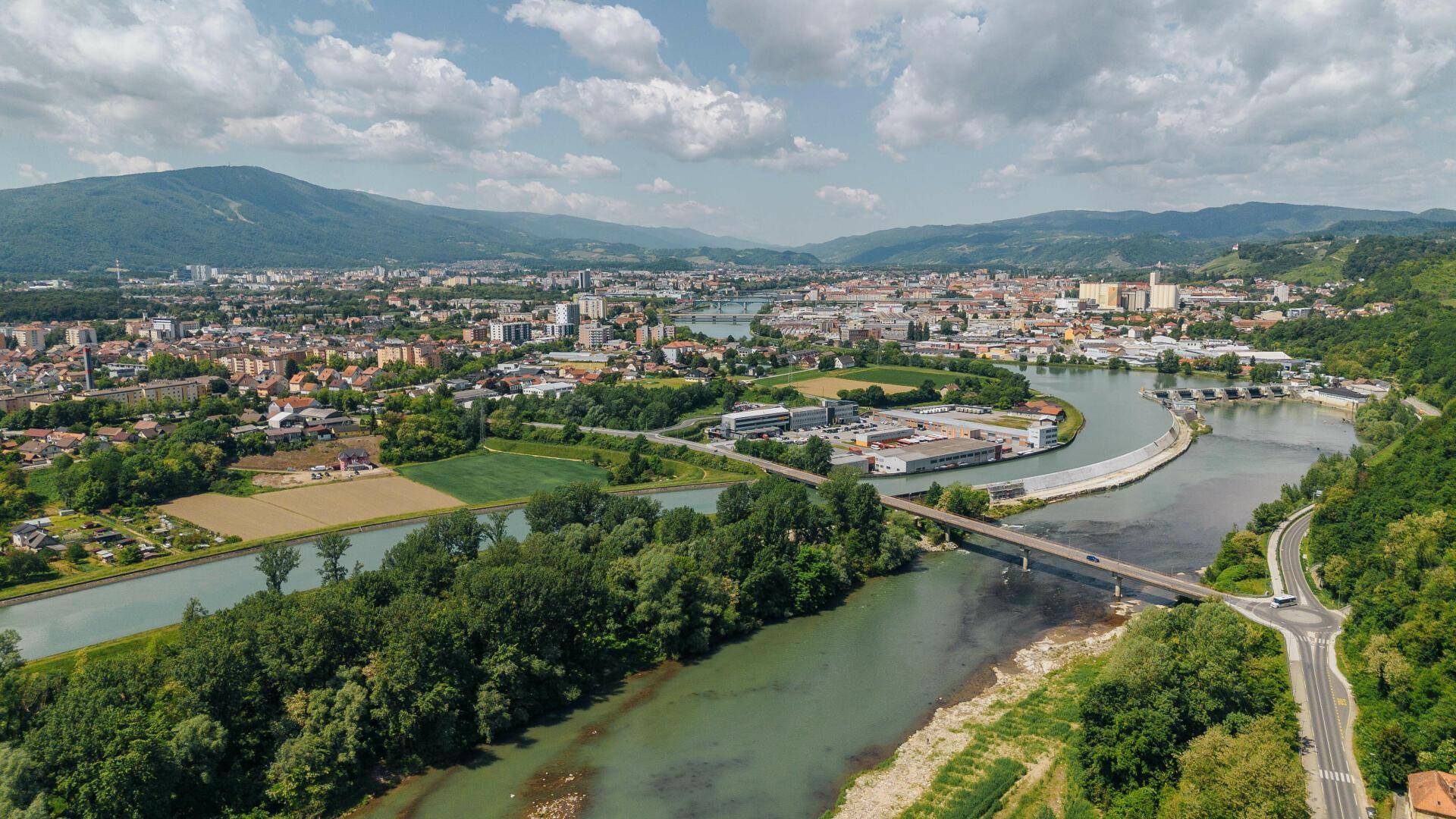 Weitläufige Luftaufnahme einer Stadt am Fluss mit Brücken und bewaldeten Hügeln im Hintergrund.