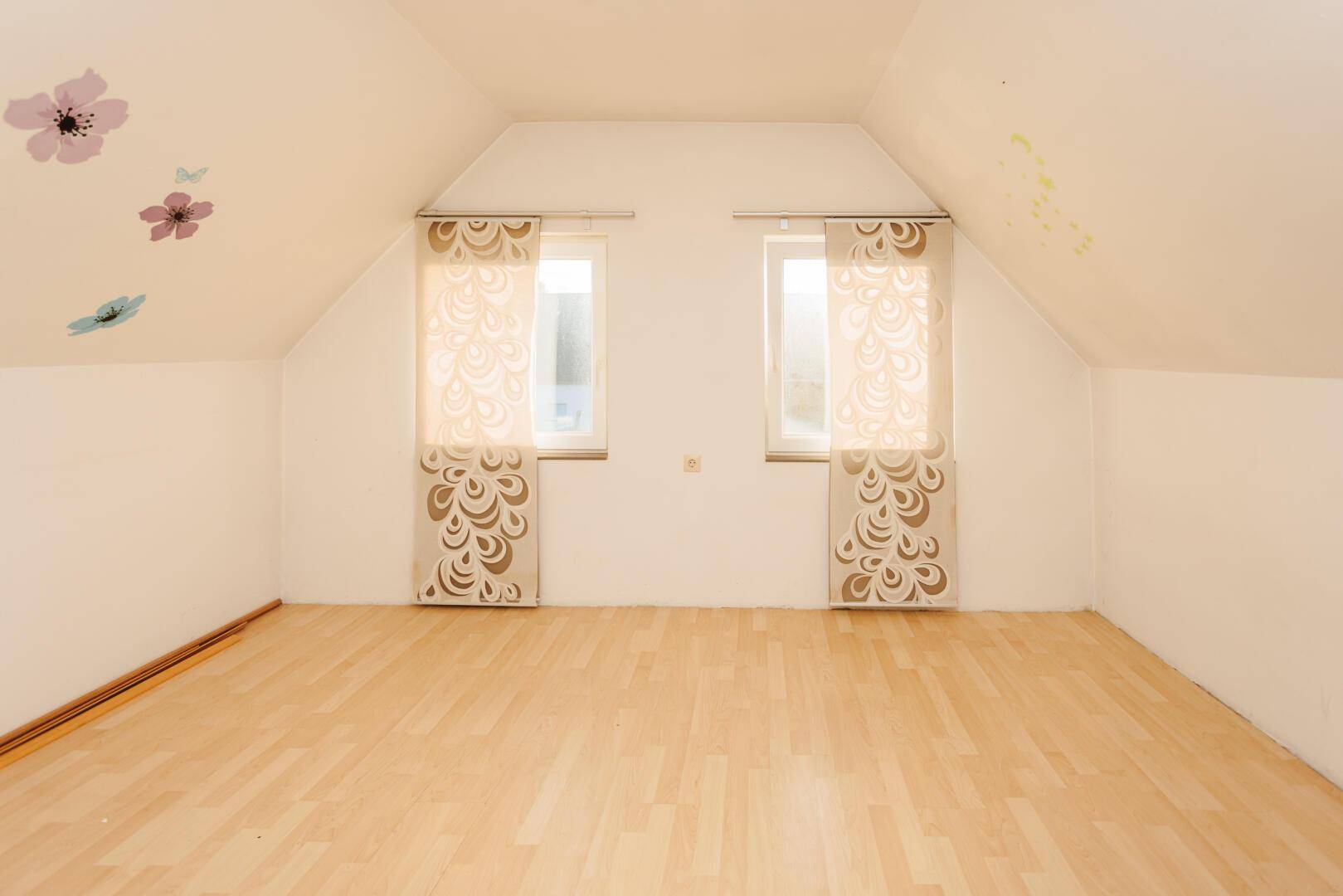 Heller Raum im Dachgeschoss mit schrägen Wänden und zwei Fenstern, dekoriert mit Blumenstickern.