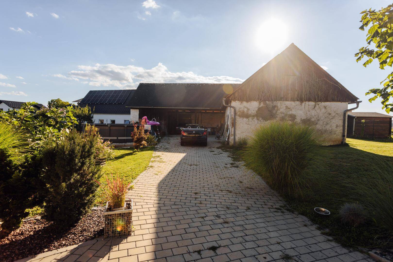 Gepflasterte Einfahrt führt zu Nebengebäuden und einem Carport, umgeben von Grünflächen.