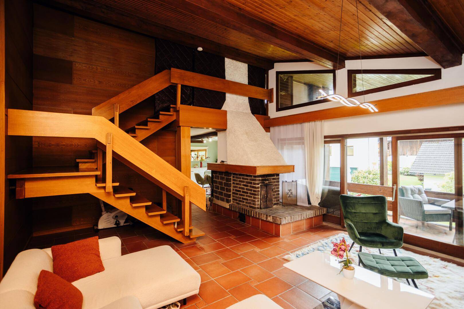 Geräumiges Wohnzimmer mit offener Holztreppe, gemauertem Kamin und Terrakottafliesen.