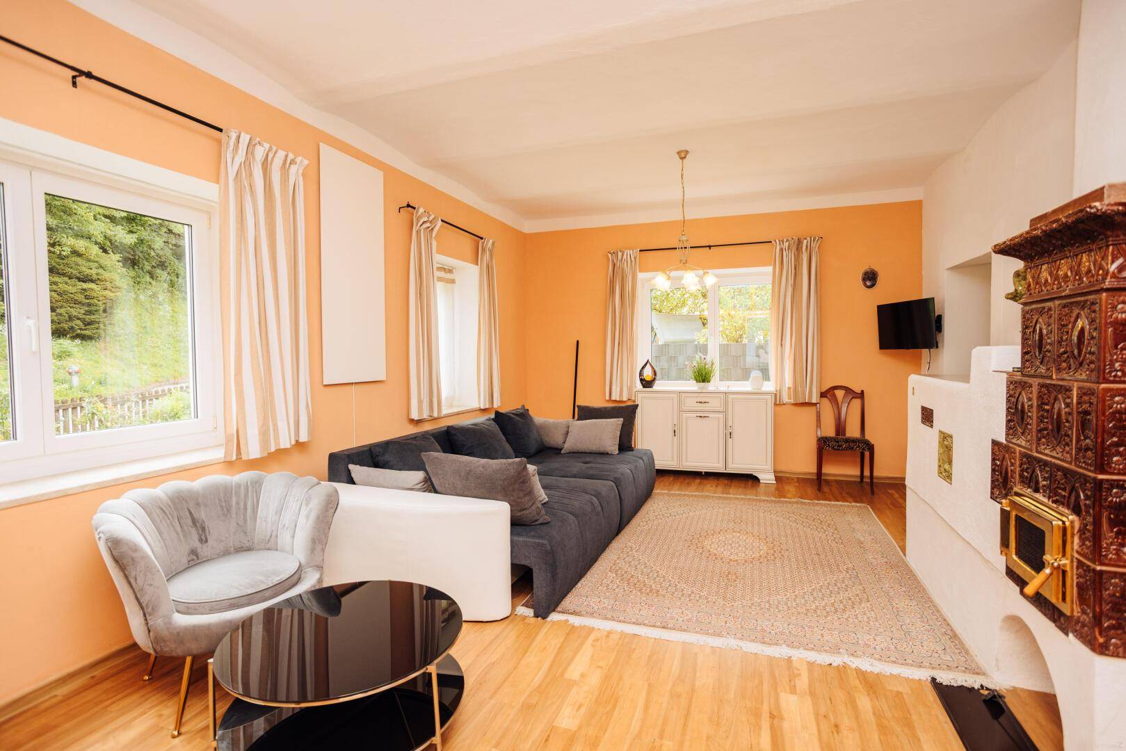 Geräumiges Wohnzimmer mit Kachelofen, bequemer Sitzecke und großen Fenstern für viel Tageslicht.