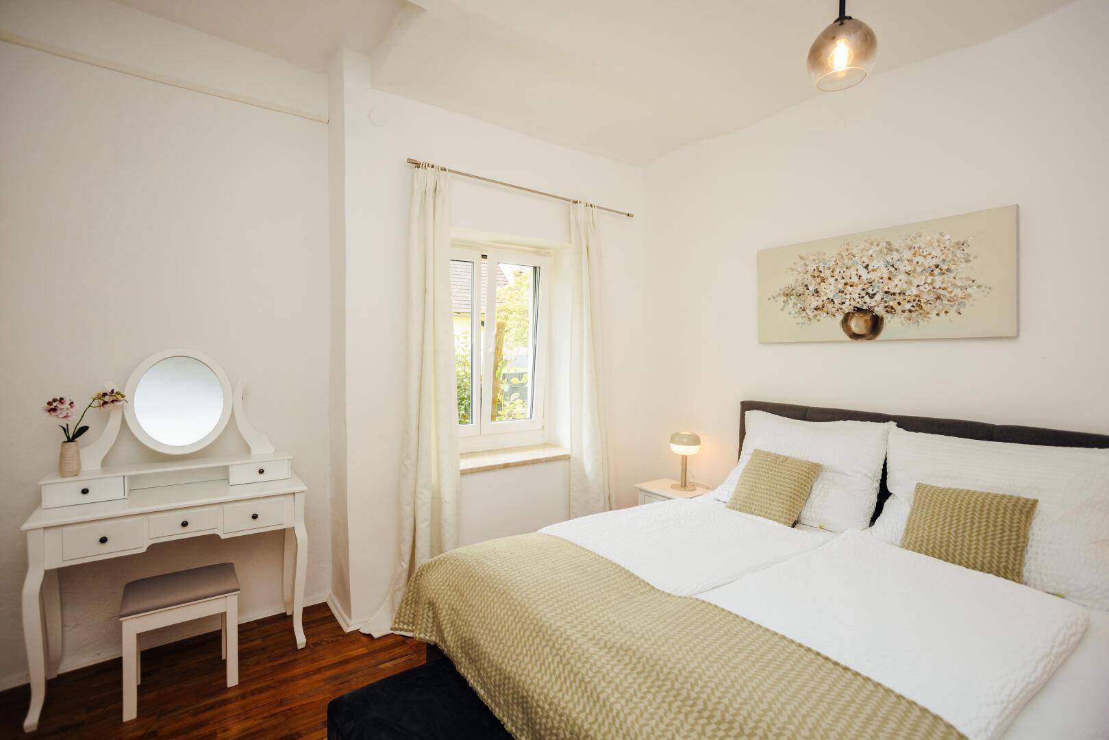 Helles Schlafzimmer mit weißem Schminktisch, Doppelbett und Blick ins Grüne durch das Fenster.