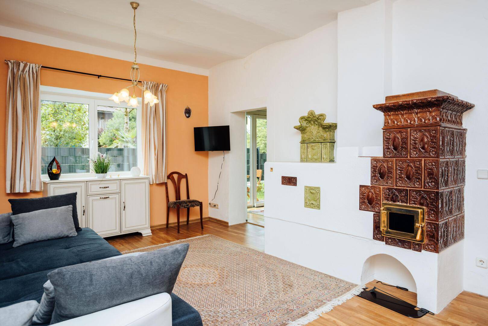 Gemütliches Wohnzimmer mit orangefarbener Wand, traditionellem Kachelofen und Holzboden.