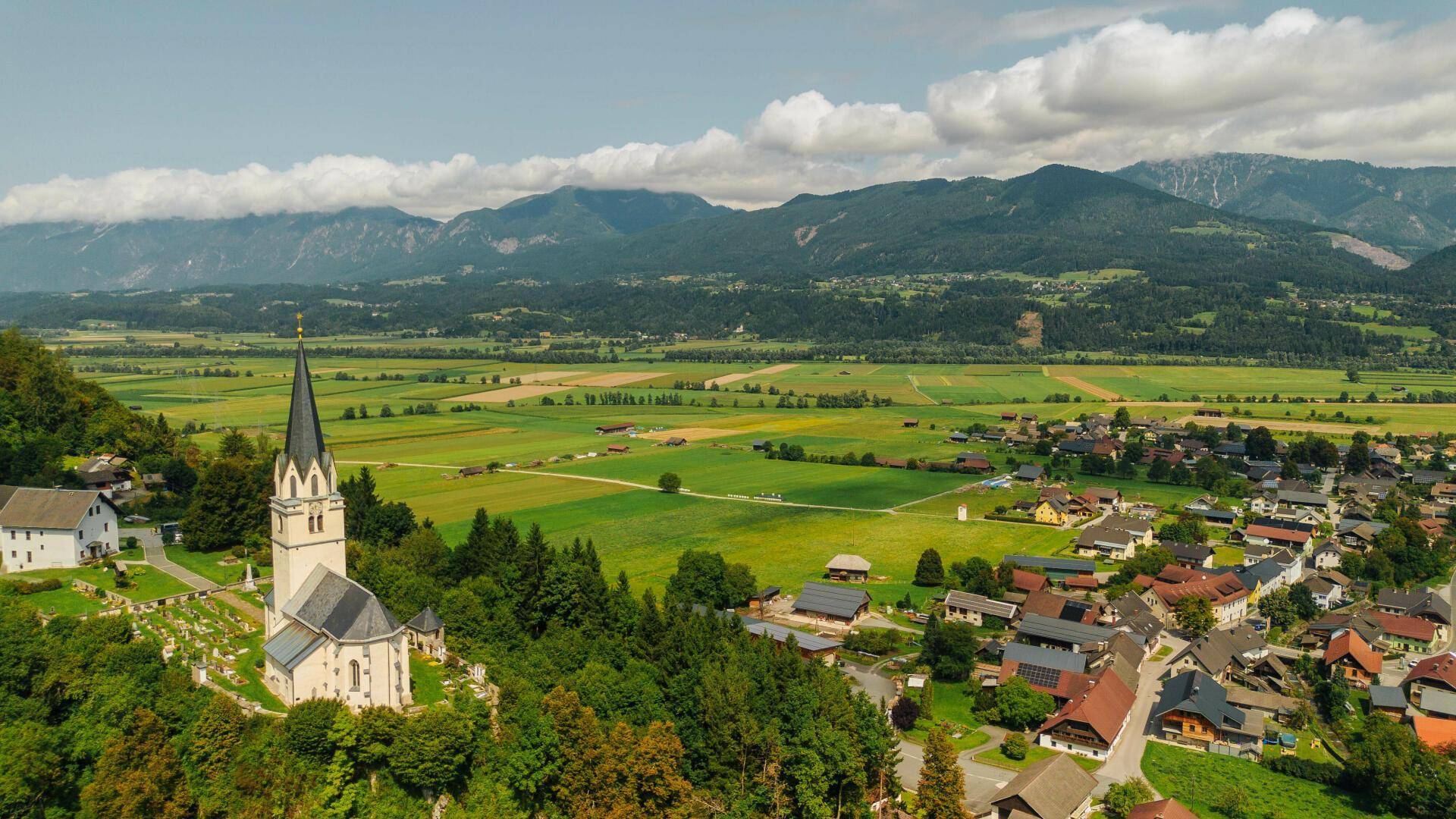 Weitläufige Luftaufnahme eines Dorfes mit Kirche, Feldern und majestätischen Bergen im Hintergrund unter blauem Himmel.