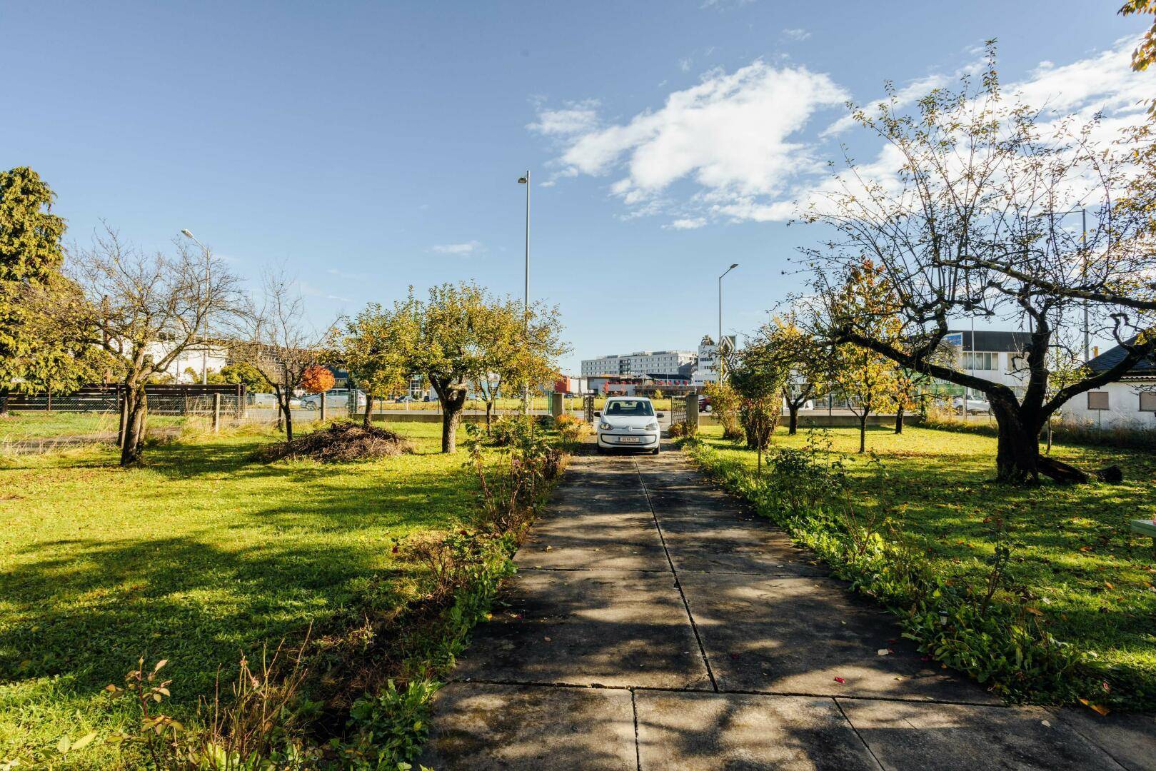 Lange Zufahrt zum Haus, umgeben von weitläufigen Grünflächen und Bäumen unter blauem Himmel.