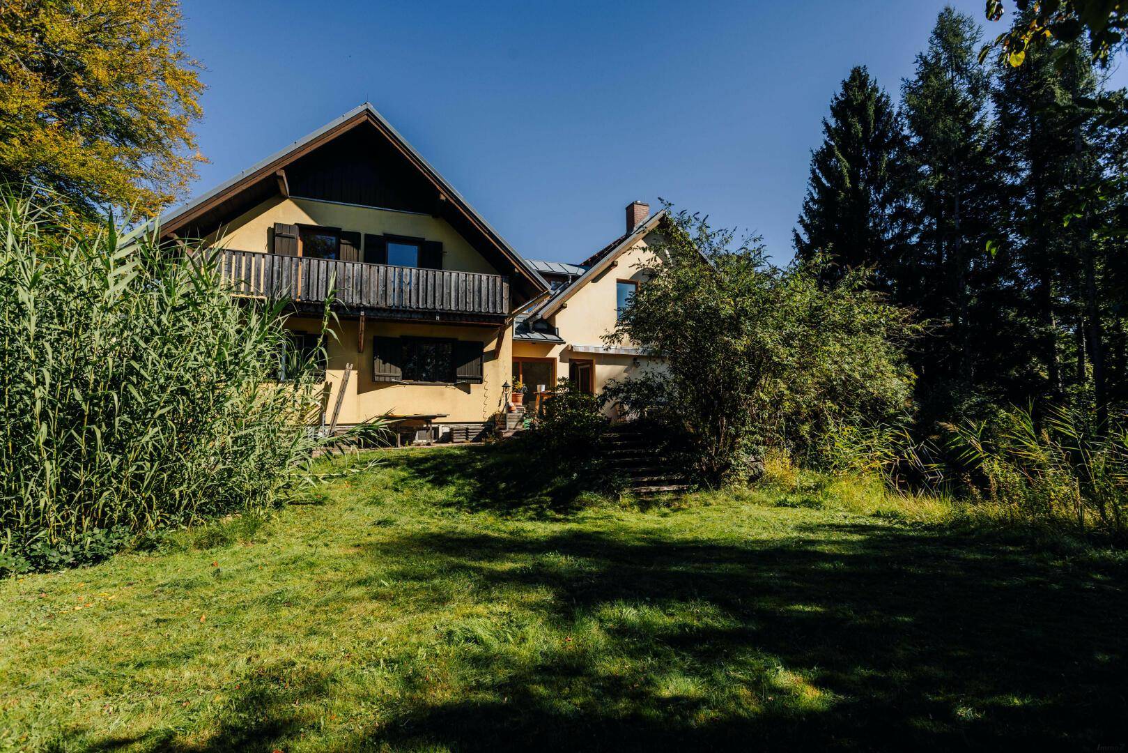 Ländliches Haus mit traditioneller Architektur, umgeben von viel Grün und Bäumen unter blauem Himmel.