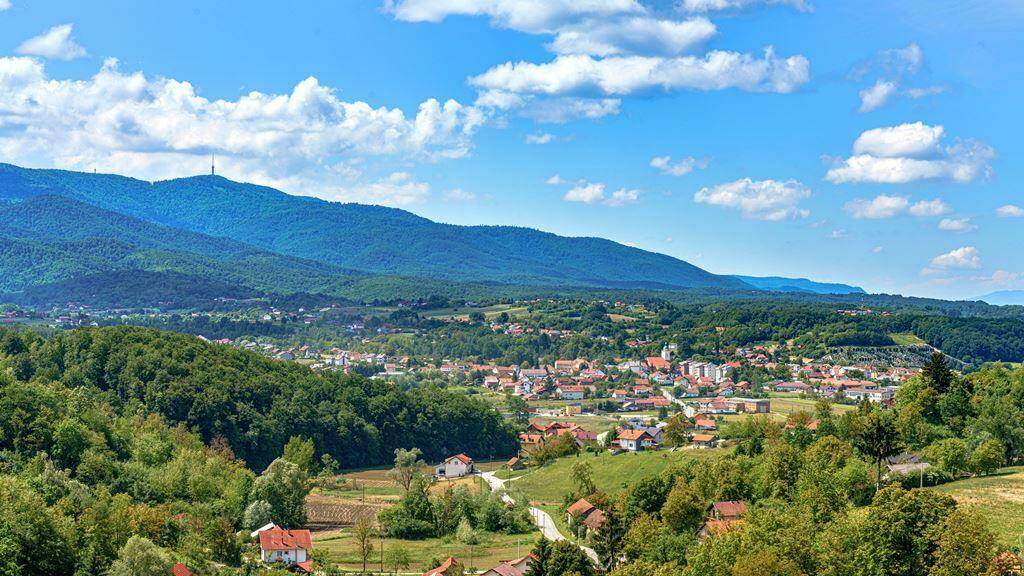 Panoramablick auf eine malerische Stadt in einem grünen Tal, umgeben von bewaldeten Hügeln unter blauem Himmel.