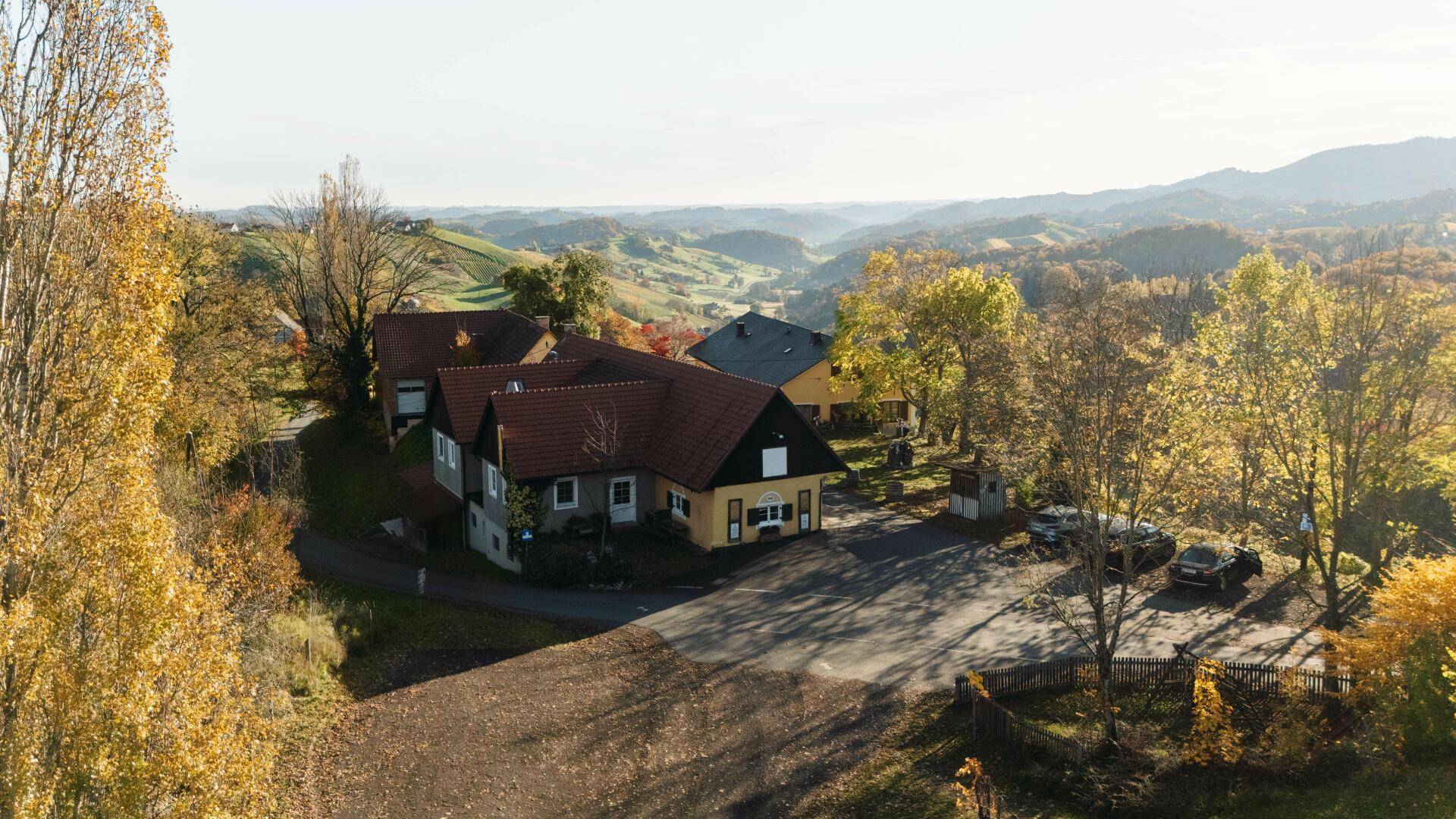 Luftaufnahme eines Anwesens mit mehreren Gebäuden und Parkplatz, umgeben von herbstlicher Landschaft und Weinbergen.